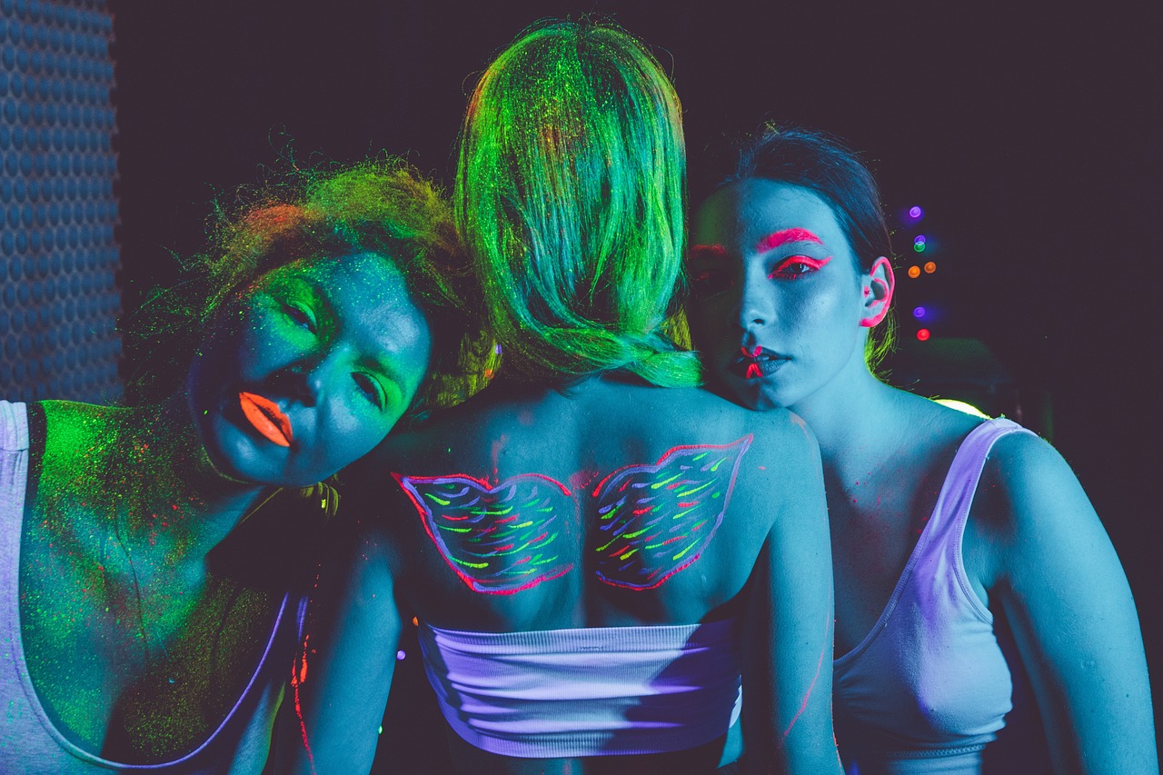 Zdjęcie przedstawia trzy młode kobiety, środkowa odwrócona jest plecami do oglądającego, atmosfera zdjęcia przypomina zdjęcie klubowe, z imprezy w stylu techno